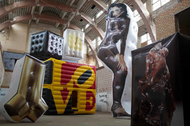 large inflatable art installation featuring oversized printed sculptures including a tall female figure, cubes and abstract shapes inside an industrial warehouse